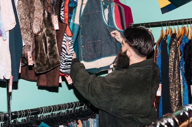A person browsing through racks of clothing at a thrift store, with a focus on the selection of vintage jackets, graphic tees, and unique streetwear accessories.