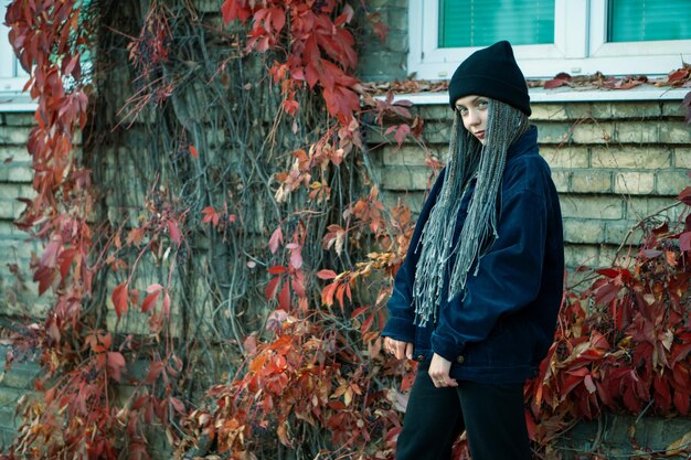 A woman wearing an oversized grey hoodie layered under a black leather bomber jacket, paired with ripped jeans and combat boots. She is wearing a beanie and a scarf.