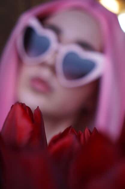 A close-up shot of a person wearing oversized, geometric-shaped sunglasses with a translucent neon pink frame. The background is blurred, focusing on the reflective lenses and the wearer's bold, confident expression.