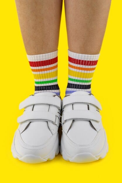 A close-up of a person wearing chunky white sneakers paired with brightly colored, patterned socks that peek out from the top of the shoes. The socks feature bold stripes and quirky graphic designs, adding a playful contrast to the clean, minimalist sneakers.