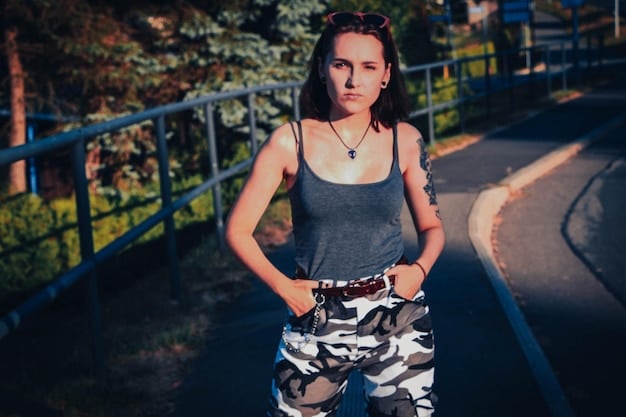 A woman in a casual women's streetwear outfit, sporting a camouflage jacket and a white t-shirt. She has a colorful bandana tied loosely around her neck, enhancing the outfit's laid-back vibe and complementing her simple makeup. The setting is an urban park.