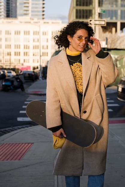 A street style photograph featuring a person wearing a tan trench coat layered over a gray sweater and black pants. They are accessorized with a colorful scarf and fashionable sunglasses.