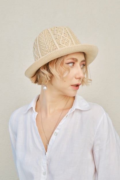 A detailed shot focusing on the head of a styled woman wearing a neutral-colored bucket hat with subtle embroidery, paired with oversized silver hoop earrings and a clean makeup look. The background is a slightly blurred urban wall.