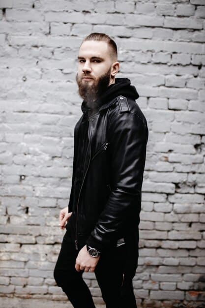 A man with a short beard, wearing a black leather jacket and a layered set of silver chains, one with a prominent pendant, against a blurred city lights background at night. The jewelry is detailed and visible.