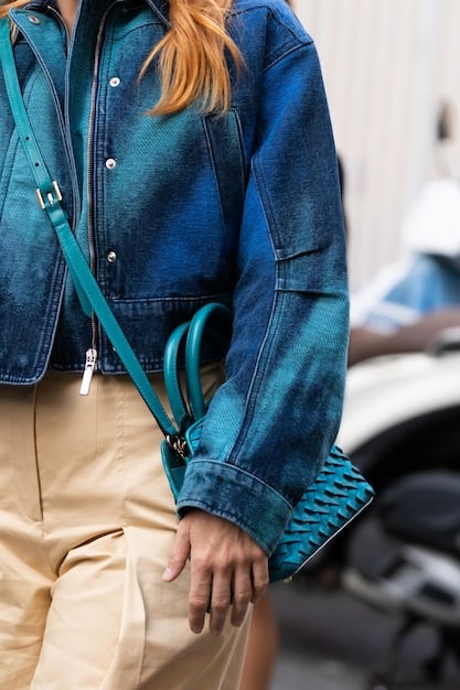 A close-up shot of a minimalist black leather crossbody bag, with a sleek metal buckle, worn over a denim jacket. The background is slightly blurred with urban elements like street art.